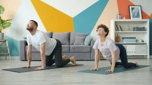 A photo of two people performing the cat cow stretch on yoga mats in a living room.